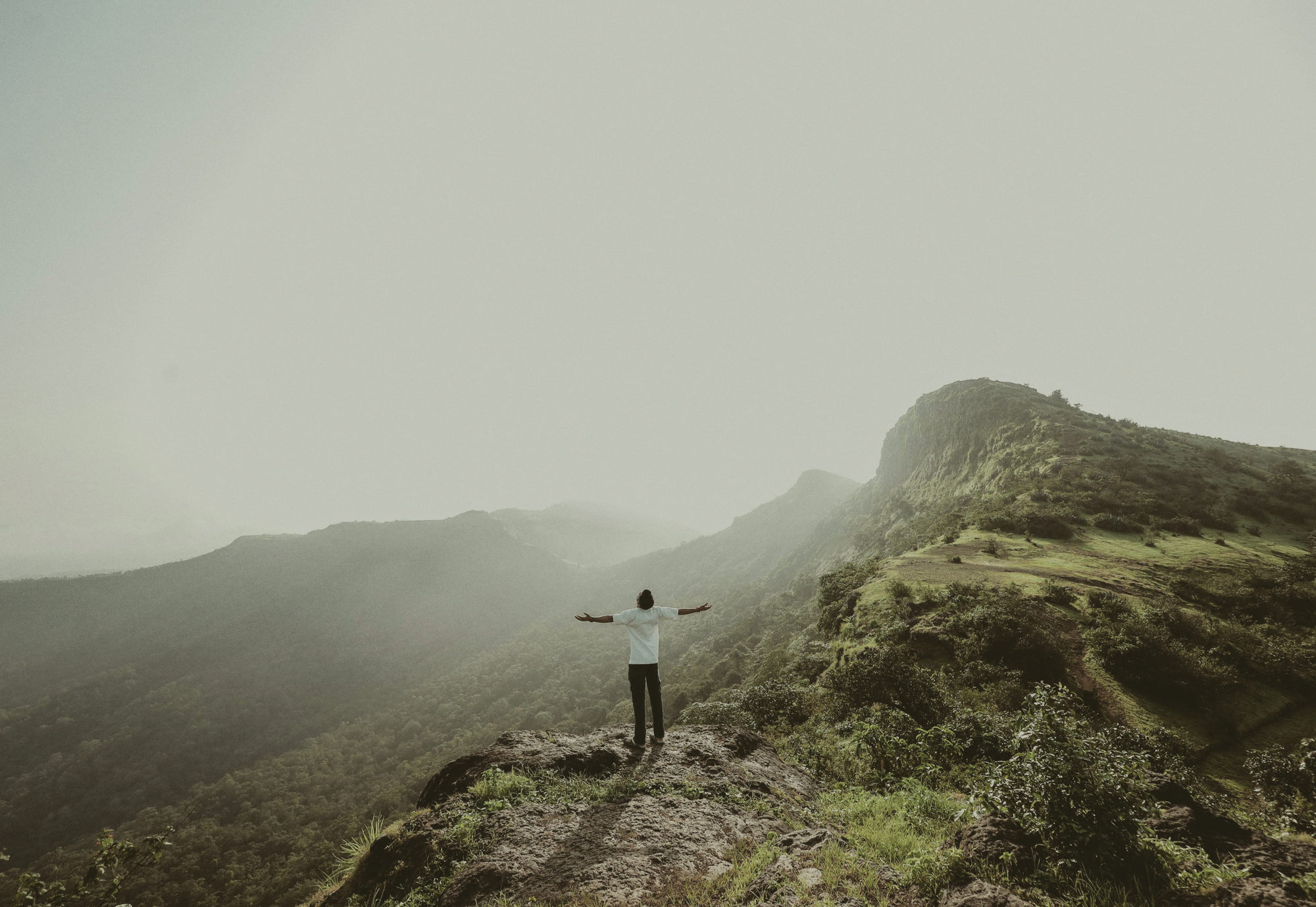 Person standing on a mountain ridge with arms outstretched, overlooking misty green mountains.